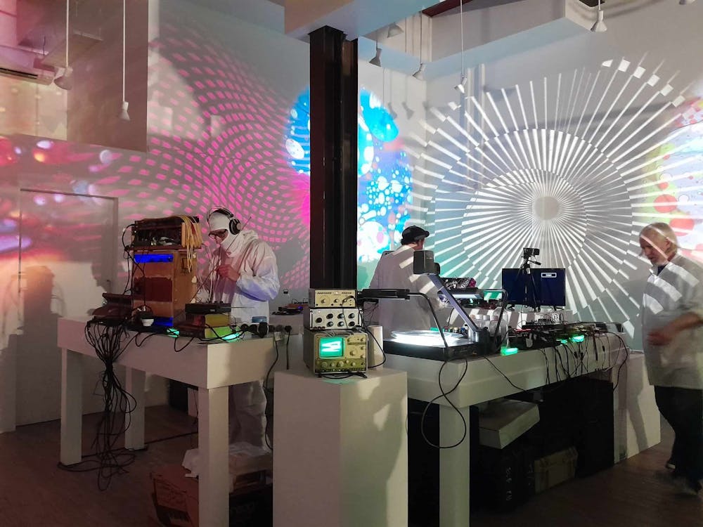 Men in white coats playing analogue synthesizers with lights lit up in psychadelic colours.