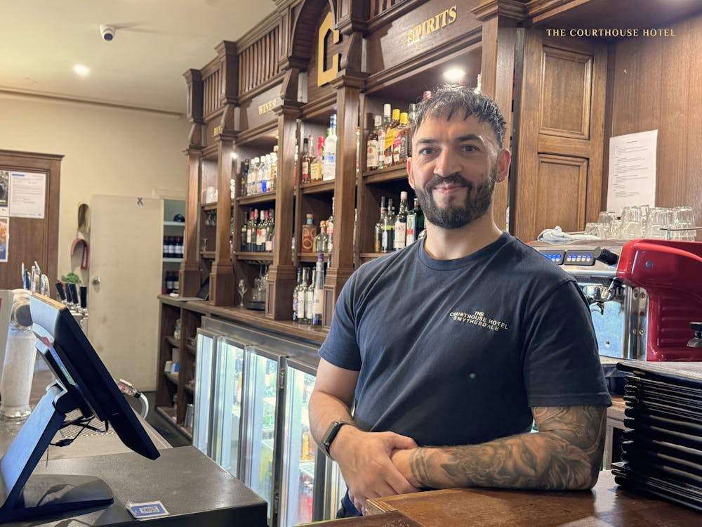 A staff member standing behind the bar at the Courthouse Hotel with shelves of spirits in the backgr