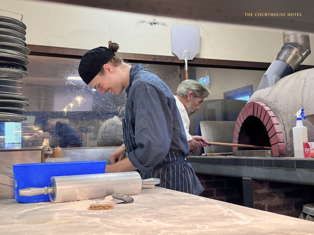 A chef preparing pizza dough on a bench in the Courthouse Hotel kitchen with a pizza oven in the bac