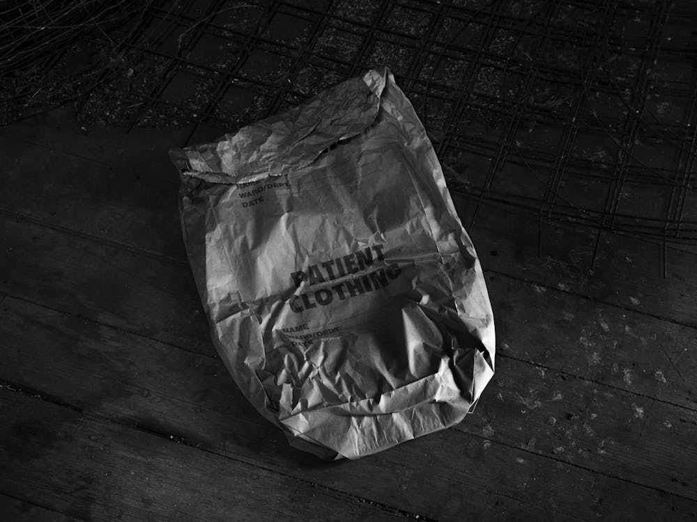 Black and white photograph of a crumpled hospital patient clothing bag on a wooden floor.
