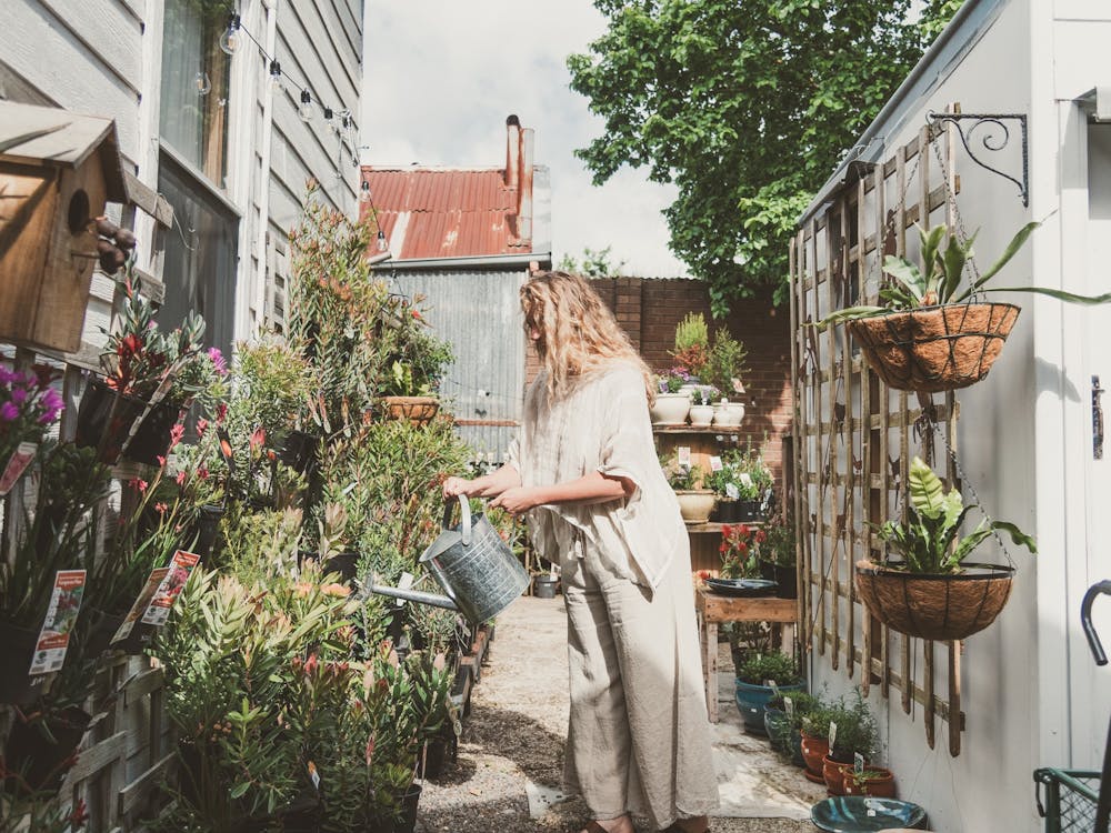 Woman wearing white linen outfit, watering plants with tin watering can in a plant nursery.