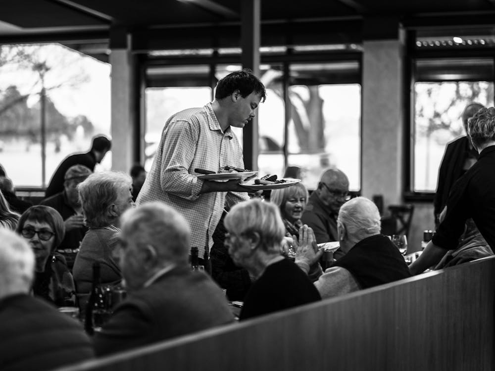 Staff member serving meals in a busy dining room filled with guests enjoying dinner together.