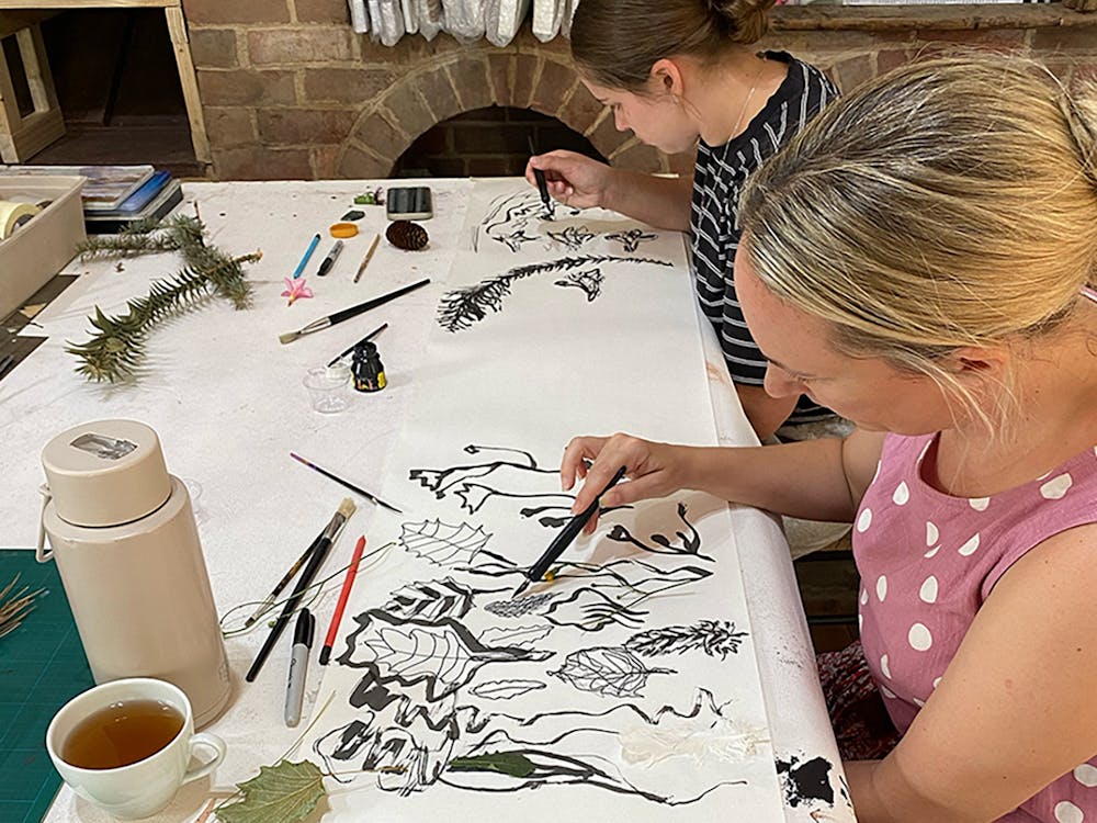 Two people drawing botanical forms in ink at a workshop table, historic brick fireplace behind.