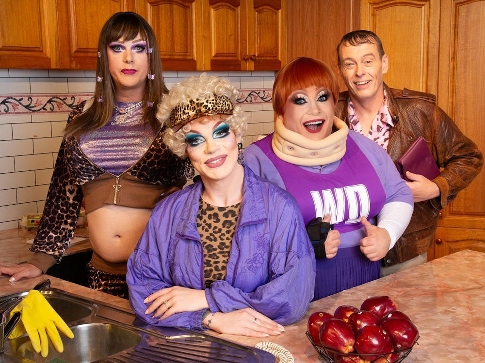Four performers in colourful Y2K-inspired costumes and theatrical makeup pose together in a kitchen,