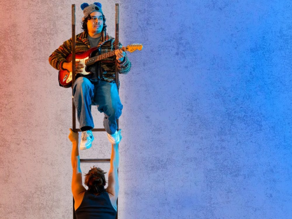 A musician sits at the top of a ladder playing a guitar. The ladder is being held upright in the air