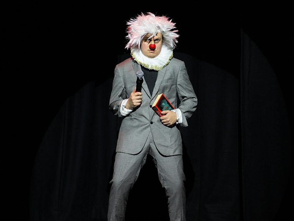 Artist in grey suit with white afro wig , red nose and white frilly color
