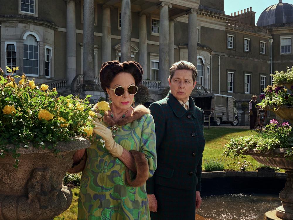 Two women stand in manor house garden next to yellow roses